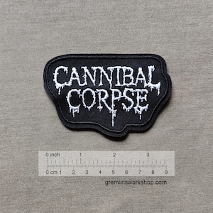 May include: Black embroidered patch with the text "CANNIBAL CORPSE" in white, featuring a dripping effect. The patch is approximately 3 inches wide. The background is a neutral tan color. The website gremlinsworkshop.com is visible.