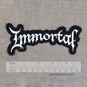Könnte beinhalten: Schwarzer Aufnäher mit dem Wort "Immortaf" in weißer gotischer Schrift. Der Aufnäher ist etwa 10 cm lang. Der Aufnäher befindet sich auf einem grauen Stoff. Die Website gremlinsworkshop.com ist sichtbar.