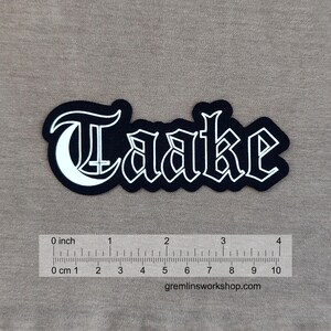 May include: Black patch with the word "Taake" in white gothic lettering. The patch is approximately 10 cm long. The website gremlinsworkshop.com is printed at the bottom.