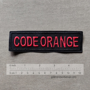 Könnte beinhalten: Schwarzer, rechteckiger Aufnäher mit dem rot gestickten Schriftzug "CODE ORANGE". Der Aufnäher ist etwa 10 cm lang. Ein Lineal darunter zeigt Messungen in Zoll und Zentimetern.