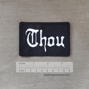 Könnte beinhalten: Schwarzer gestickter Aufnäher mit dem Wort "Thou" in weißen Buchstaben. Der Aufnäher ist etwa 5 cm breit und 2,5 cm hoch.