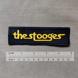 Op de afbeelding: Een rechthoekige, marineblauwe geborduurde patch met de gele tekst "the stooges". De patch is ongeveer 10 cm lang. Een liniaal is eronder geplaatst.