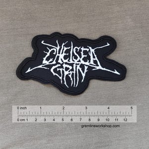 Könnte beinhalten: Schwarzer, gestickter Aufnäher mit dem weißen Text "CHELSEA GRIN" in einer stilisierten Schriftart. Der Aufnäher hat eine unregelmäßige Form und einen schwarzen Rand. Der Aufnäher ist etwa 12 cm breit, wie das Lineal zeigt.