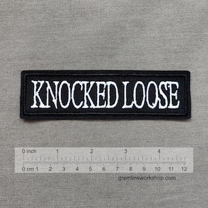 Puede incluir: Parche rectangular negro con el texto "KNOCKED LOOSE" bordado en blanco. El parche mide aproximadamente 10 cm de largo. El fondo es de tela gris neutra.
