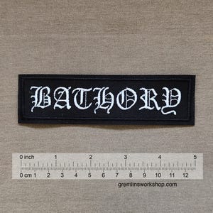 May include: A rectangular black embroidered patch featuring the white text BATHORY in a gothic font. A ruler placed below the patch indicates a length of approximately 4.5 inches. The patch has a stitched border and a flat, fabric texture.