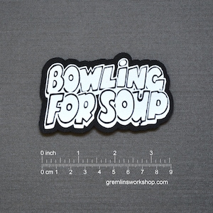 May include: A black and white patch with the text "BOWLING FOR SOUP" in a bubble font. The patch has a black border and is set against a gray background. The bottom of the image has a ruler with inches and centimeters.
