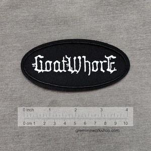 Puede incluir: Parche ovalado negro con el texto blanco "GoatWhore" en fuente gótica. El parche mide aproximadamente 10 cm de largo. El fondo es una tela gris.