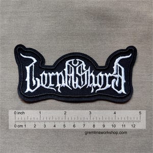 Op de afbeelding: Zwarte geborduurde patch met het woord "Lorphidhora" in witte gotische letters. De patch is ongeveer 12,7 cm lang en heeft een unieke vorm met gebogen randen. De patch ligt op een bruinachtige achtergrond.