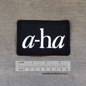 Può includere: Una toppa rettangolare nera con la scritta bianca "a-ha" ricamata. La toppa ha un bordo nero ed è posta su uno sfondo grigio. È visibile un righello con pollici e centimetri.