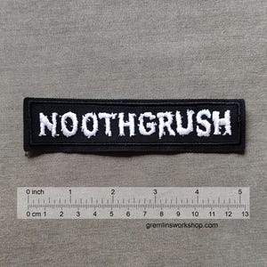 Könnte beinhalten: Schwarzer rechteckiger Aufnäher mit dem gestickten Wort "NOOTHGRUSH" in Weiß. Der Aufnäher ist etwa 13 cm lang. Der Hintergrund ist ein neutraler grauer Stoff. Die Website gremlinsworkshop.com ist sichtbar.