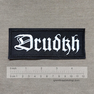 May include: Black rectangular patch with the word "Drudkh" embroidered in white gothic lettering. The patch measures approximately 10 cm in length. The background is a neutral tan colour.