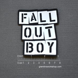 Puede incluir: Un parche rectangular con las palabras "FALL OUT BOY" en letras blancas en negrita, estilo plantilla, sobre fondos negros. El parche mide aproximadamente 7.6 cm de ancho. El fondo es una tela gris lisa.