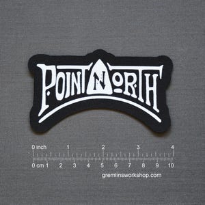 Könnte beinhalten: Schwarzer und weißer Aufnäher mit dem Text "POINT NORTH" in einer stilisierten Schriftart. Der Aufnäher hat eine gebogene Unterkante und ein kleines Dreieck über dem Buchstaben "N". Der Aufnäher ist ca. 10 cm breit.