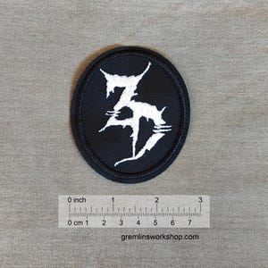 Könnte beinhalten: Schwarzer und weißer gestickter Patch mit dem Buchstaben "Z" in einer stilisierten Schriftart. Der Patch ist oval geformt und hat einen Durchmesser von etwa 5 cm.