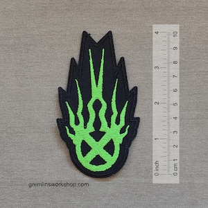May include: Black patch with a green flame design, approximately 7.6 cm tall. The patch features a stylized flame pattern with a central circular design. The website gremlinsworkshop.com is visible at the bottom.
