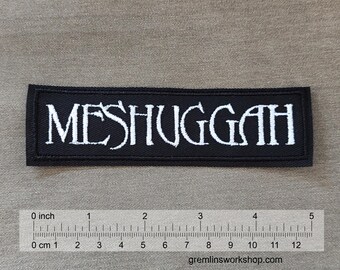 Może przedstawiać: Czarno-biała naszywka haftowana z napisem "MESHUGGAH" w białych literach na czarnym tle. Naszywka ma około 10 cm szerokości.