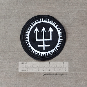 Könnte beinhalten: Ein runder, schwarzer Aufnäher mit einem weißen Dreizack-Symbol in einem weißen Sonnenmuster. Der Aufnäher hat einen Durchmesser von etwa 7.6 cm. Die Website gremlinsworkshop.com ist unten aufgedruckt.