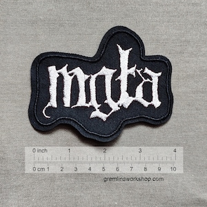 Pode incluir: Etiqueta bordada preta com a palavra "mata" em letras brancas. A etiqueta tem aproximadamente 10 cm de largura e 7,5 cm de altura.