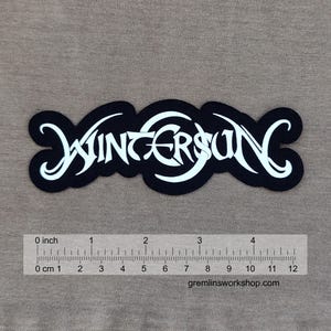May include: Black and white patch with the word "Wintersun" in a decorative font. The patch measures approximately 10 cm in length. The patch is displayed on a neutral background.