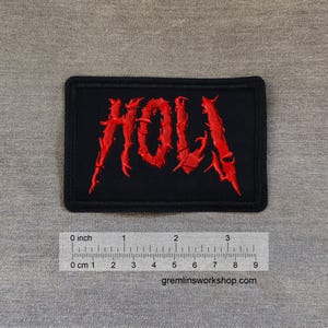May include: A rectangular black patch with the word "HOLA" embroidered in red, stylized with sharp, jagged edges. The patch includes a ruler with inch and centimeter measurements, and the website gremlinsworkshop.com.