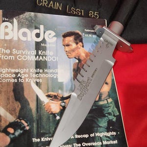 May include: A silver survival knife with a black handle, resting on a magazine cover featuring a man holding a knife. The magazine is titled "The Blade" and has the text "The Survival Knife From COMMANDO!". The knife is labeled "CRAIN LSS1 85".