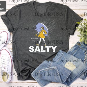 May include: Dark heather gray t-shirt with a graphic of a person under an umbrella in the rain, with the word "SALTY" in white. The shirt is paired with blue jeans and white sneakers.