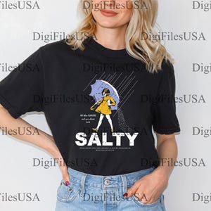 May include: Black t-shirt featuring a graphic of a person under an umbrella, with the word "SALTY" in bold white letters. The person is wearing a yellow dress and holding a blue umbrella. The shirt has the text "All the flavors and you choose to be".