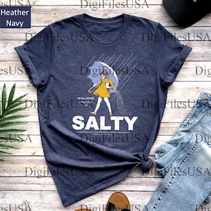May include: Heather navy t-shirt with a graphic of a person under an umbrella, with the word "SALTY" in white. The person is wearing a yellow coat. The shirt is paired with blue jeans.
