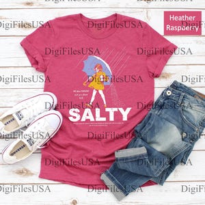 May include: Heather raspberry t-shirt with a graphic of a person under an umbrella, with the word "SALTY" in white. The shirt is paired with blue jeans and white sneakers.