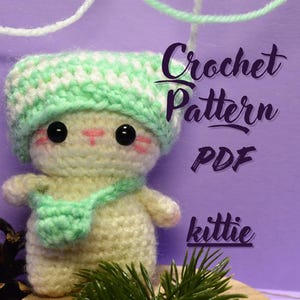 May include: Crochet pattern PDF for a white and green kitty amigurumi toy. The kitty is wearing a green and white striped hat and a green scarf. The text "Crochet Pattern PDF" and "kittie" are visible in the image.