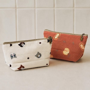 Può includere: Due pochette con cerniera in tessuto. La pochette anteriore è color bianco sporco con volti di gatto e impronte di zampe ricamati. La pochette posteriore è color arancio terracotta con motivi di arachidi e zucche. Tirazip a forma di gatto.