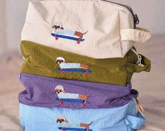 May include: A stack of four rectangular zippered pouches made of grid-patterned fabric in cream, olive green, purple, and light blue. Each pouch features an embroidered dachshund wearing a white helmet and striped shirt on a blue skateboard.