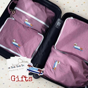Op de afbeelding: Een set mauvekleurige packing cubes met ruitpatroon en witte ritsen, elk voorzien van een geborduurde teckel op een blauw skateboard. Een witte kaart met de tekst Travel Packing List Gifts en een teckel-sleutelhanger zijn inbegrepen.