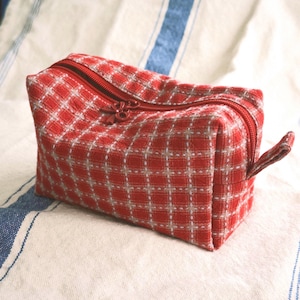 Puede incluir: Un estuche rectangular de tela con un patrón de cuadros rojos y blancos. El estuche tiene una cremallera roja en la parte superior con un pequeño tirador decorativo. La tela tiene una apariencia texturizada y una pequeña trabilla lateral.