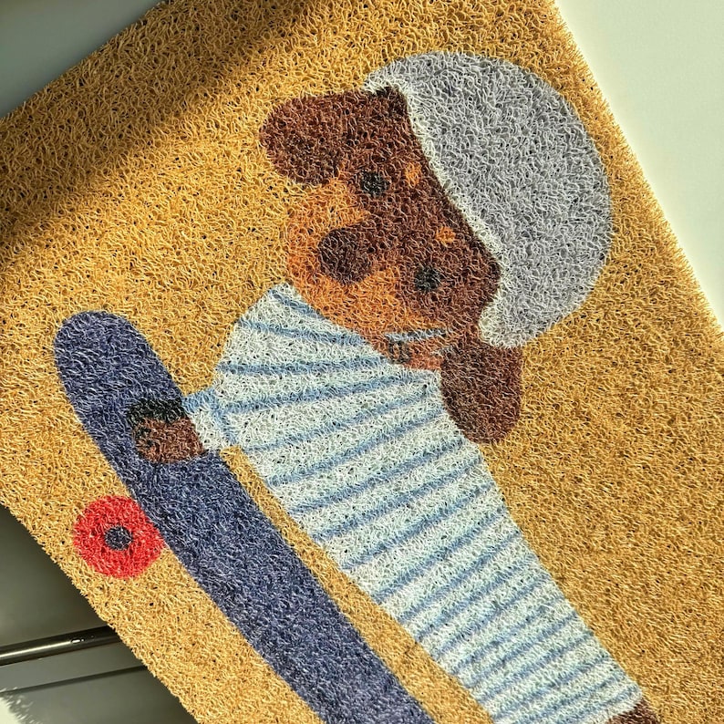 May include: A rectangular doormat featuring a cartoon dog riding a skateboard. The dog has brown fur and a gray helmet, wearing a blue and white striped shirt. The skateboard is blue with a red wheel, set against a tan background.
