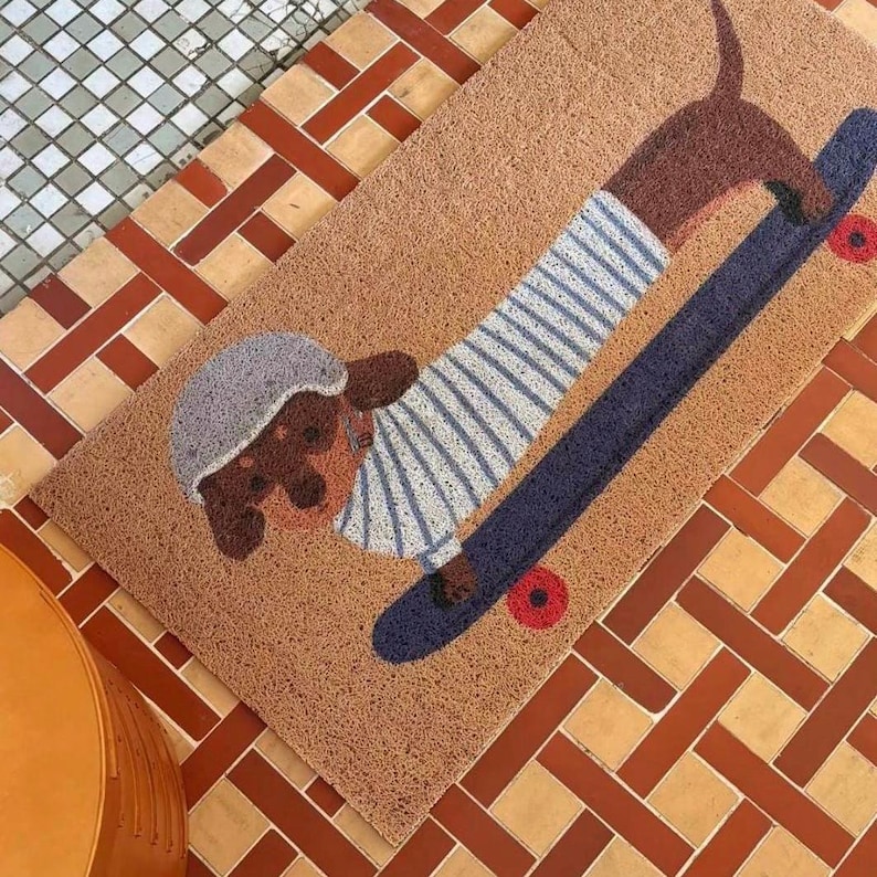 May include: A rectangular doormat featuring a brown dachshund dog on a skateboard. The dog wears a striped shirt and a gray helmet. The skateboard is navy blue with red wheels. The mat is brown with a textured surface.