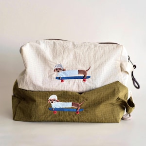 May include: Two rectangular zippered pouches, one cream and one olive green, both featuring a grid pattern. Each pouch displays an embroidered graphic of a brown dachshund wearing a white helmet and blue striped shirt, riding a blue skateboard.