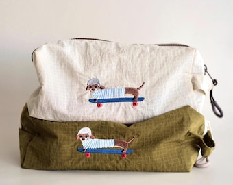 May include: Two zippered pouches, one off-white and one olive green, both featuring a grid pattern. Each pouch displays an embroidered graphic of a brown dachshund wearing a white helmet and a light blue striped shirt while riding a blue skateboard.