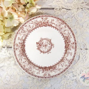 Pu&ograve; includere: Un piatto in ceramica bianca con un motivo floreale marrone dettagliato. Il piatto ha un design floreale centrale e un bordo decorativo. Il piatto &egrave; su una superficie di pizzo bianco con fiori gialli sullo sfondo.