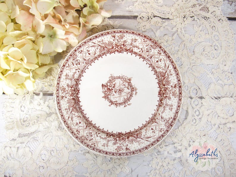 Pu&ograve; includere: Un piatto in ceramica bianca con un motivo floreale marrone dettagliato. Il piatto ha un design floreale centrale e un bordo decorativo. Il piatto &egrave; su una superficie di pizzo bianco con fiori gialli sullo sfondo.