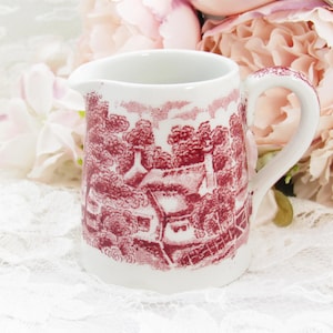 May include: A small, white ceramic creamer with a red toile pattern depicting a rural landscape. The creamer has a handle and a spout for easy pouring. The background features soft pink flowers.