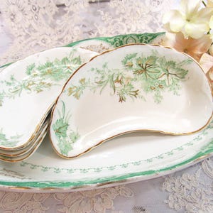 May include: A collection of antique white porcelain serving dishes with a green floral design and gold trim. The set includes several kidney-shaped dishes and a large oval platter, all arranged on a lace tablecloth.