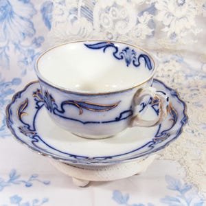 May include: A white teacup and saucer set with blue floral designs and gold trim. The cup and saucer are placed on a small, white, three-legged stand. The background features a floral patterned fabric.