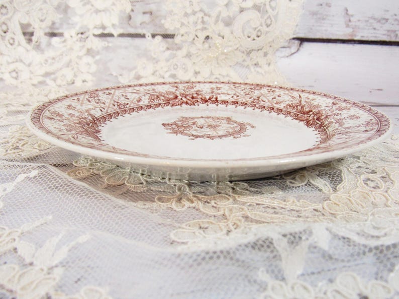 Pu&ograve; includere: Un piatto in ceramica bianca con un dettagliato motivo floreale e decorativo marrone sul bordo. Il piatto ha un motivo a medaglione centrale ed &egrave; appoggiato su una tovaglia di pizzo. Un piatto decorativo in stile vintage.