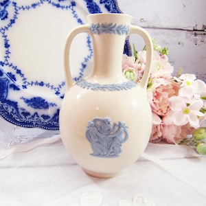 May include: A white ceramic vase with two handles and a blue and white floral design. The vase features a raised design of three figures in blue.