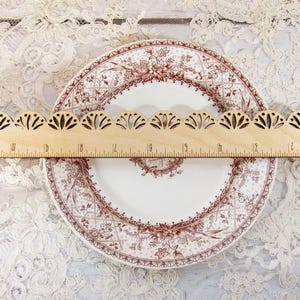 Pu&ograve; includere: Un righello di legno con un bordo smerlato decorativo &egrave; appoggiato su un piatto vintage. Il piatto ha uno sfondo bianco con un disegno floreale marrone. Il righello &egrave; contrassegnato con misure in centimetri.