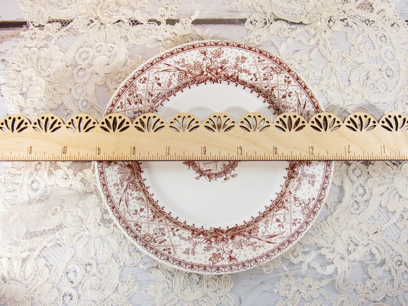 Pu&ograve; includere: Un righello di legno con un bordo smerlato decorativo &egrave; appoggiato su un piatto vintage. Il piatto ha uno sfondo bianco con un disegno floreale marrone. Il righello &egrave; contrassegnato con misure in centimetri.
