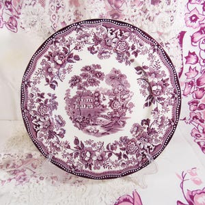 May include: A white plate with a purple floral and landscape pattern. The plate has a scalloped edge and a dark purple dotted border. The central design shows a building, bridge, and trees. The plate is a classic design.
