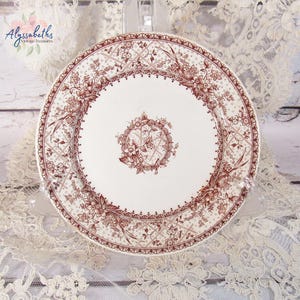 Pu&ograve; includere: Un piatto in ceramica bianca con un motivo floreale marrone dettagliato. Il piatto presenta un medaglione floreale centrale e un bordo decorativo. Il piatto &egrave; esposto su una tovaglia di pizzo, mostrando un'estetica vintage.