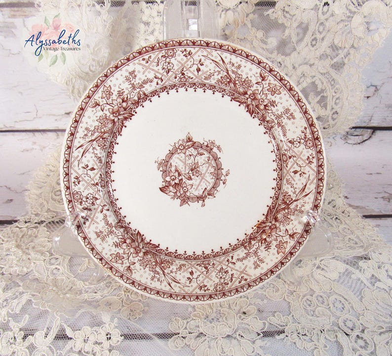 Pu&ograve; includere: Un piatto in ceramica bianca con un motivo floreale marrone dettagliato. Il piatto presenta un medaglione floreale centrale e un bordo decorativo. Il piatto &egrave; esposto su una tovaglia di pizzo, mostrando un'estetica vintage.